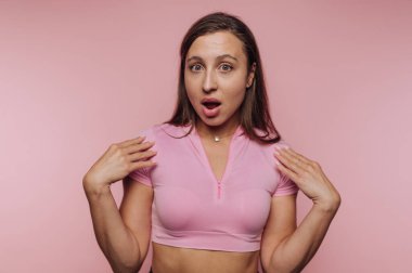 Genç bir kadın pembe bir arkaplanda duruyor, şaşırdığını ve heyecanlandığını ifade ediyor. Pembe bir bluz giyer ve duygularını canlandırıcı bir hareketle gösterir..