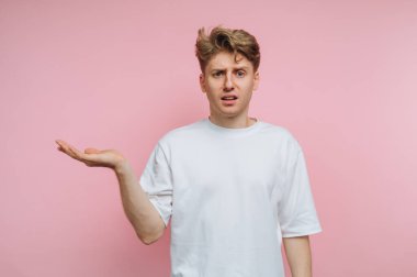 Kafası karışmış beyaz bir tişört giymiş genç bir adam sorgu jesti için elini kaldırıyor. Yumuşak pembe arka plan neşeli bir atmosfer yaratır..