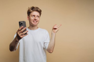 Genç bir adam akıllı bir telefon tutuyor ve kameraya gülümsüyor. Diğer eliyle hareket ediyor, heyecanlı ya da hevesli görünüyor. Arka plan basit ve tarafsız..