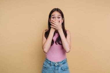 Bir kız şaşırmış, elleri kameranın dışında bir şeye şaşkın şaşkın bakarken ağzını kapatıyor. Pembe bir bluz ve kot şort giyiyor, neşeli bir atmosferi çağrıştırıyor..