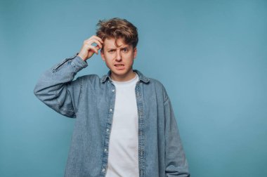 Genç bir adam koyu mavi bir arkaplan önünde duruyor, kafası karışmış ve kaşınıyor, bir kafa karışıklığı yaşıyor ve çözülmemiş bir sorun hakkında derin düşünceler taşıyor..