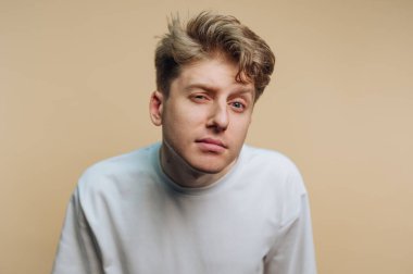 Saçları dağınık genç bir adam, sağlam bir arkaplan önünde bir gözünü kısarak belirsizlik gösteriyor. Düz beyaz bir gömlek giyiyor, rahat ve samimi bir görünüm sergiliyor..