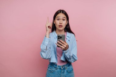 Uzun siyah saçlı genç bir kadın akıllı telefonunu tutarken şaşırmış görünüyor. Bir parmağını kaldırmış, pembe bir arka plana yaslanmış bir şekilde el kol hareketi yapıyor. Bu da bir şeyin farkına varma ya da keşfetme anı olduğunu gösteriyor..