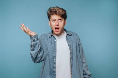 Açık kahverengi saçlı genç bir adam eliyle el kol hareketi yaparken düş kırıklığı belirtileri gösteriyor. Düz mavi bir fonda duruyor, beyaz bir gömleğin üzerine kot ceket giyiyor..
