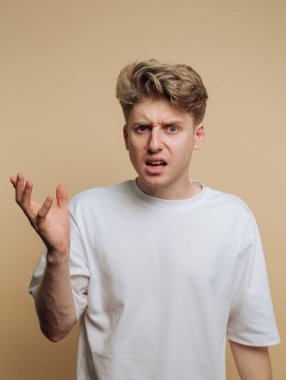 Genç bir adam yüzünde şaşkın bir ifadeyle duruyor, tek eliyle işaret ediyor. Basit bir kapalı mekan ortamında kafa karışıklığı veya hayal kırıklığını ifade ediyor gibi görünüyor..