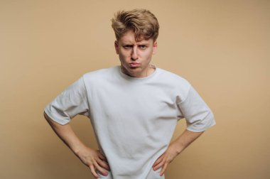 Saçları dağınık genç bir adam elleri kalçasında duruyor, somurtuyor ve hafif, nötr bir tonda sade bir arka plana karşı komik bir ifade sergiliyor..