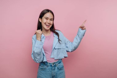 Genç bir kadın açık mavi gömlek ve kot şort giymiş canlı pembe bir arka plana gülümsüyor. Heyecanını baş parmak ve işaret hareketleriyle belli ediyor..