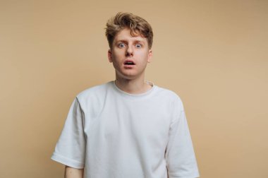 Genç bir adam, yüzünde şok olmuş bir ifadeyle sıradan bir tişörte şaşkınlık ve güvensizliği yansıtan sade bir arkaplan karşısında duruyor..