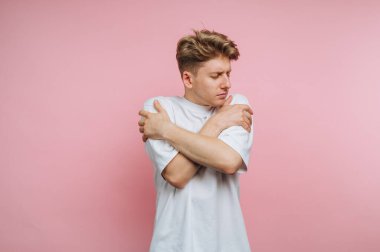 Genç bir adam, kollarını kendi etrafına sarmış, gözleri kapalı, yumuşak pembe bir arka fonun önünde dikilirken yansıyor. Onun günlük kıyafetleri rahat atmosfere katkı sağlar..