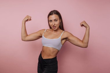 Genç bir kadın pembe bir zemine karşı poz veriyor, kaslarını göstermek için gururla kollarını esnetiyor. Kendine güveni ve gücü yayar, fitness yoluyla güçlendirme gücü verir..