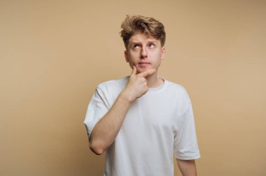 Genç bir adam eli çenesinde duruyor, dalgın dalgın bakıyor. Günlük beyaz gömleği minimalist bej arka planla çelişiyor ve yansıtıcı bir atmosfer yaratıyor..