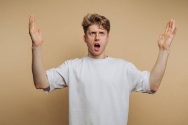 Genç bir adam açık bir ağızla ayakta duruyor ve ellerini kaldırıyor, güçlü bir şaşkınlık veya şok ifadesi sergiliyor. Arka plan sıcak, nötr bir ses tonu, tepkisini arttırıyor..