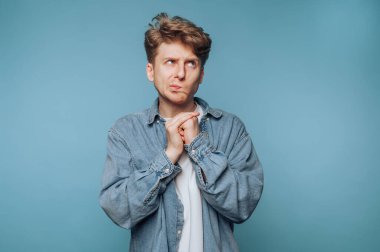 Genç bir adam mavi bir arkaplan önünde duruyor, bir kot gömlek ve beyaz tişört giyiyor, düşünceli bir görünüm ve bir soru ya da fikir üzerine kafa yorar gibi ellerini sıkarak.