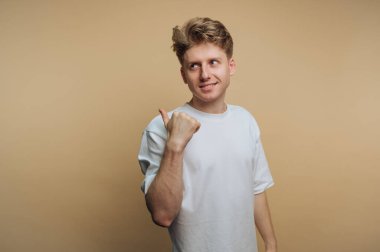 Açık renk saçlı genç bir adam rahat bir iç mekanda başparmağını arkasını göstererek güvenle gülümsüyor. Tarafsız bir arkaplanda düz beyaz bir tişört giyiyor..