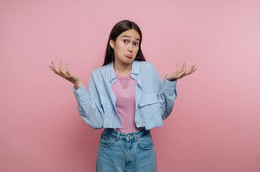 Genç bir kadın pembe bir arka planın önünde duruyor, omuzlarını silkiyor ve kafası karışmış bir ifade sergiliyor. Pembe bluz ve kot pantolon üzerine açık mavi çizgili bir gömlek giyer..