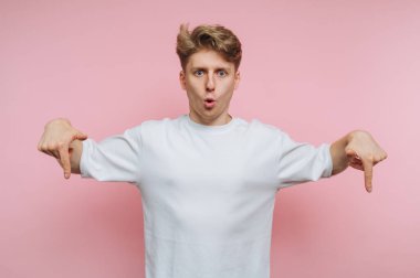 Genç bir adam sürpriz bir ifadeyle pembe bir zemine karşı duruyor, ağzı açık ve iki eliyle aşağıya bakıyor. Onun gündelik, rahat kıyafeti neşe katıyor..