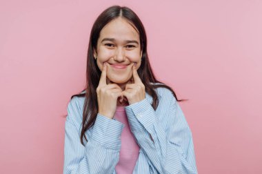 Bir kız neşeli pozlar veriyor, parmakları yanaklarında neşeli bir hareketle gülümsüyor. Pembe arka plan onun neşeli yüz ifadesini geliştiren canlı bir atmosfer yaratıyor..