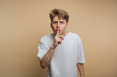 Genç bir adam parmaklarını dudaklarına dayayıp sessizliği işaret ediyor. Saçı dağınık, gözleri açık, sade beyaz bir gömlek giymiş, neşeli bir merak yayıyor..