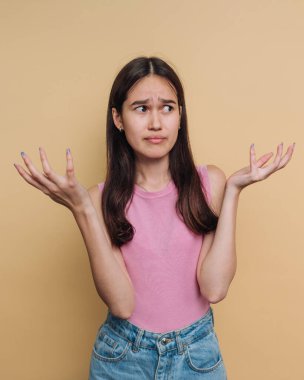 Şaşkın bir ifadeyle genç bir kadın sorgulama jesti için elini kaldırıyor. Pembe bir bluz ve kot pantolon giyiyor, sıcak, tarafsız bir arka plan üzerine kurulmuş..