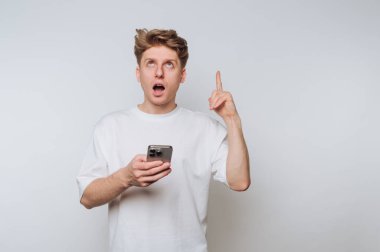 Beyaz tişört giyen genç bir adam akıllı telefonunu tutarken şaşırmış bir şekilde yukarı bakıyor. Daha yeni farkına varmışa benziyor. Parlak ışık içerideki atmosferi güçlendirir..