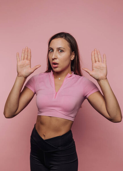 A young woman with an athletic build stands against a solid pink background, raising her hands in an expression of surprise. She wears a short-sleeved pink top and black pants.