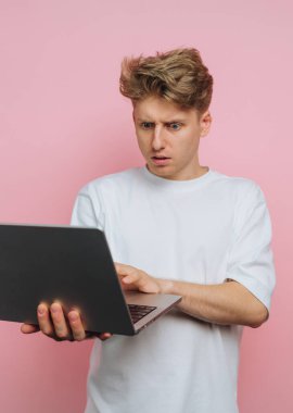 Beyaz tişörtlü genç bir adam dizüstü bilgisayarına bakarken şaşırmış bir ifade sergiliyor. Arka plan, bu beklenmedik anda gösterdiği dramatik tepkiyi artıran yumuşak bir pembe renktir..