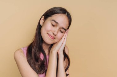 Neşeli genç bir kadın huzur içinde gülümsüyor, başını ellerinin üzerinde dinlendiriyor. Rahatlamış duruşu ve içerik ifadesi, mutluluk saçan sıcak sarı bir zemine karşı bir huzur anını vurguluyor..