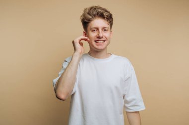 Açık kahverengi saçlı, gülümseyen ve tek eliyle el sallayan genç bir adam, kapalı mekan ortamında dostane bir tavır sergiliyor. Tarafsız zemin sıcak bir atmosfer yaratır..