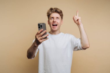 Genç bir adam, bir parmağını kaldırmış, akıllı bir telefonu tutarken heyecanını ve neşesini ifade eder. Arkaplan bej renginde, heyecanını vurguluyor..