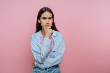 Genç bir kadın eli çenesinde açık mavi çizgili bir gömlek ve kot pantolon giyiyor. Düşünceli bir ifade sergiliyor, canlı pembe bir arkaplan üzerine kurulu..