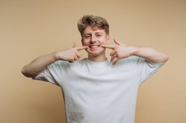 Açık kahverengi saçlı neşeli genç bir adam parlak bir gülümseme sergiliyor ve basit bir bej arka planda dişlerine şakayla işaret ediyor. Ayarlama mutluluk ve güven taşır..