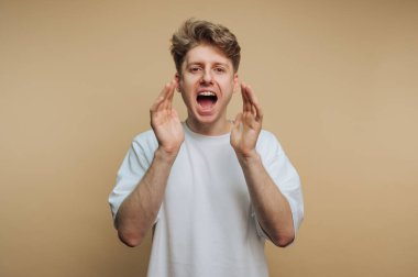 Açık kıvırcık saçlı genç bir adam tutkulu bir şekilde sevinçli bir ifadeyle bağırıyor, bej renkli bir arka planda bir anlık coşku sergiliyor..