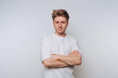 Genç bir adam kollarını tarafsız bir arkaplanda kavuşturarak güvenle ayakta duruyor. Düz beyaz bir gömlek giyiyor, stüdyo çekimi sırasında sıradan ama şık bir tavır sergiliyor..