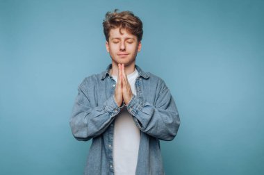 Genç bir adam meditasyon jesti olarak elleri ve gözleri kapalı bir şekilde ayakta duruyor. Sıradan giyinmiş ve açık mavi bir arka plana karşı barışçıl bir tavır sergiliyor..