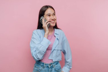 Genç bir kadın telefonuyla konuşurken gülümsüyor, pembe bir zemine karşı duruyor. Rahat ve neşeli bir hava katan sıradan bir kıyafet giyiyor..