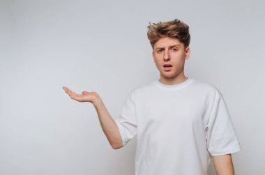 Beyaz gömlek giyen genç bir adam düz bir arkaplanda duruyor. Şaşırmış bir ifadesi ve jestleri var. Bir eli havada, kafa karışıklığı ya da inançsızlık hissi veriyor..