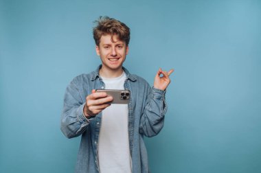 Genç bir adam, bir elinde akıllı bir telefon, diğeriyle şakalaşarak hareket ederken geniş bir gülümseme ile, sağlam bir mavi arkaplan karşısında duruyor. Gündelik kıyafetleri, canlı ortamı tamamlıyor..