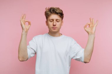 Gözleri kapalı genç bir adam farkındalık meditasyonu yapar, elleri bir Mudra jestinde, yumuşak pembe bir arka planla çevrili, sükuneti ve odaklanmayı teşvik eder..