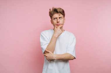 Genç bir adam eli çenesinde duruyor, düşünceli bir şekilde yana bakıyor. O, yumuşak pembe bir arka plan karşısında gündelik beyaz bir tişört giyer, bir anlık iç gözlem yansıtır.