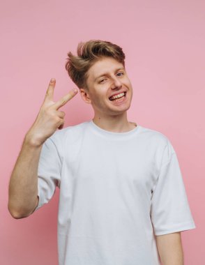Genç bir adam eliyle barış işareti gösterirken geniş bir şekilde gülümsüyor. Basit beyaz bir tişört giyiyor ve parlak pembe bir arka fonun önünde duruyor, pozitiflik saçıyor..