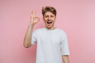 Genç bir adam neşeli bir ifadeyle duruyor, tamam işareti oluşturmak için elini kaldırıyor. Pastel pembe arka plan, neşeli tavırlarını artıran, neşeli bir his katıyor..