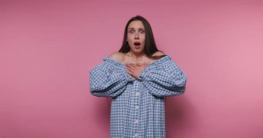 Genç bir kadının görüntüsü, göğsünü pembe arka planda sıkarken gerçek bir şaşkınlık ve şok ifadesi sergiliyor.