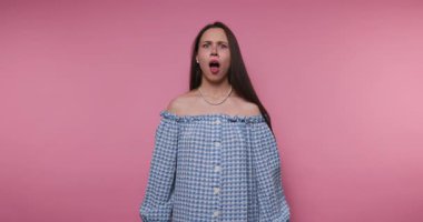 Kararlı genç bir kadının kollarını kavuşturup pembe arka planda zarif mavi kareli elbisesiyle meydan okuduğu görüntüler.