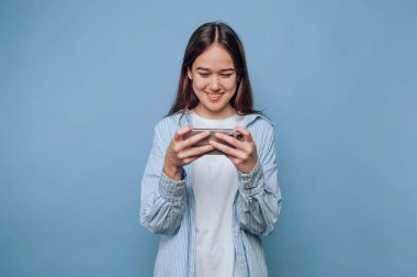 Genç bir kadın akıllı telefonuyla cep telefonu oyunu oynarken gülümsüyor. Düz mavi bir arka plana karşı duruyor, rahat ve hareketleriyle meşgul, boş zamanlarında eğleniyor..