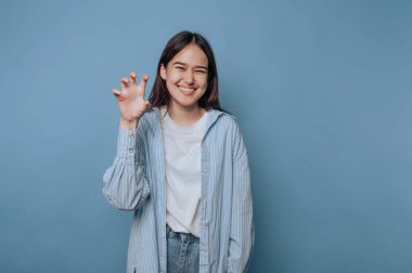 Genç bir kadın açık mavi bir ortamda eğleniyor. Çizgili bir gömlek giyer ve elleri ile neşeli bir pençe hareketi yaparken ışıl ışıl gülümser..