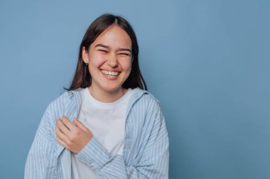 Açık mavi çizgili bir gömlek giyerken genç bir kadın parlak bir şekilde gülümsüyor. Kapalı bir alanda, yumuşak mavi bir duvara karşı neşeli bir anın tadını çıkarıyor gibi görünüyor..