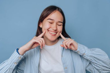 Neşeli genç bir kadın neşeyle gülümsüyor gözleri kapalı ve parmakları yanaklarını işaret ediyor. Beyaz bir gömlek ve açık mavi çizgili gömlek giyiyor. Düz mavi bir arkaplan üzerine kurulmuş..