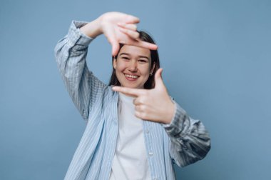 Genç bir kadın elleriyle bir çerçeve hareketi yaparken gülümsüyor. Mavi bir arka plana karşı duruyor, neşeli ve yaratıcı bir ifade sergiliyor..