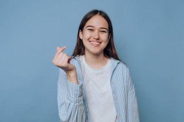 Katı mavi bir duvarın önünde duran bir kız, sağ eliyle bir parmakla kalp hareketi yaparken ışıl ışıl gülümsüyor. Sıradan beyaz bir gömlek ve açık mavi çizgili bir ceket giyiyor..