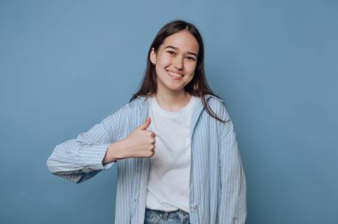 Mavi bir duvarın önünde genç bir kadın baş parmağını kaldırırken neşeyle gülümsüyor. Beyaz bir tişört ve kot şortun üzerine açık mavi çizgili bir gömlek giyiyor, pozitiflik yayıyor..